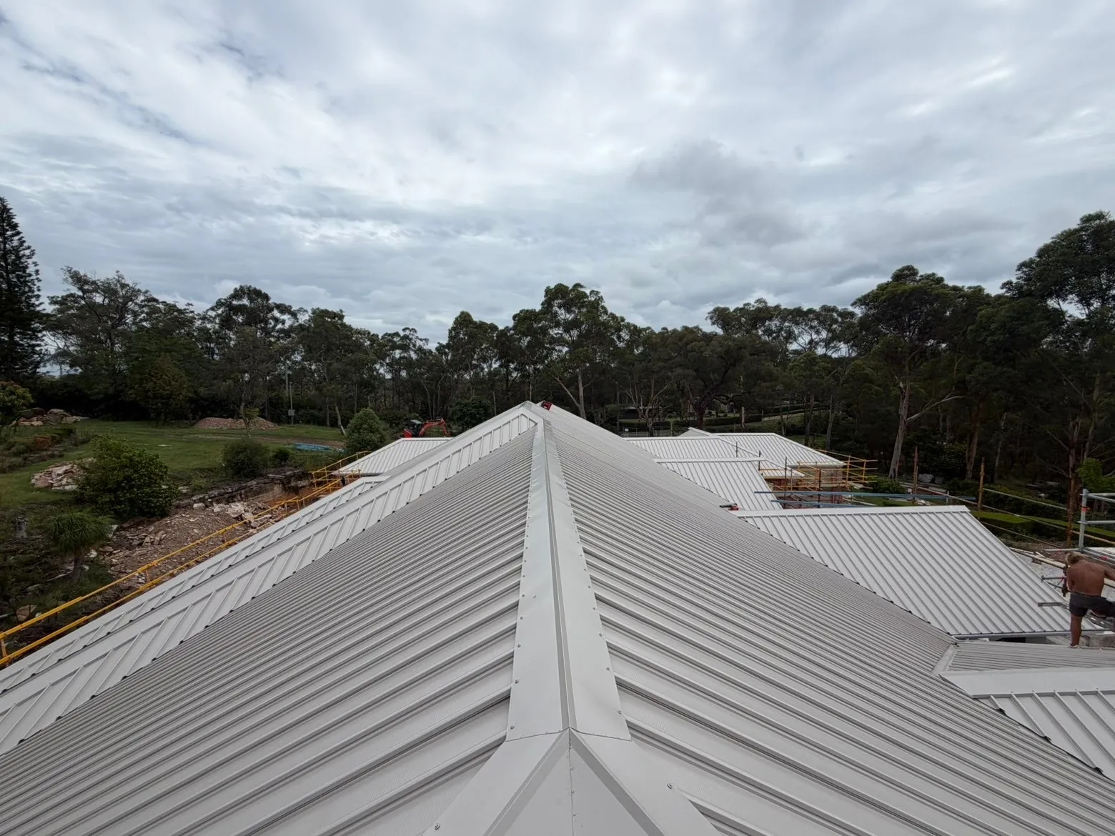 A pale ribbed metal roof on a new home in bushland, looking along a hip towards gum trees under a cloudy sky