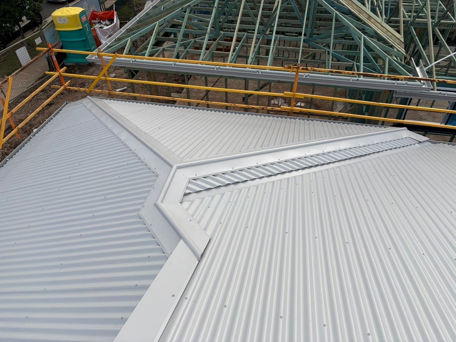 Hip, ridge and valley capping meeting on a pale corrugated metal roof, with timber roof frames on the next block