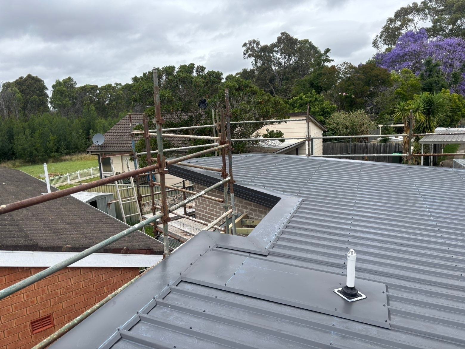 A grey ribbed metal roof with a flashed vent pipe and flashing folded along a brick wall
