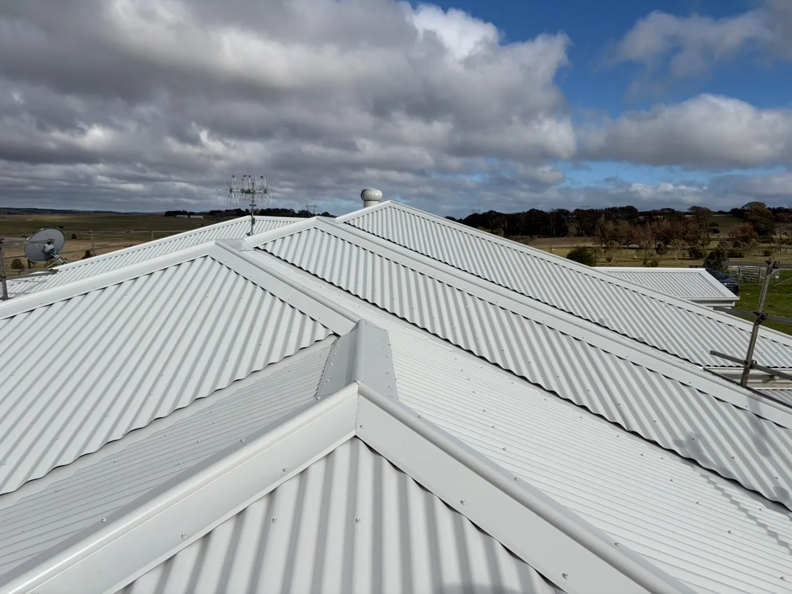 Ridge and hip capping meeting on the same pale corrugated roof, with open farmland behind