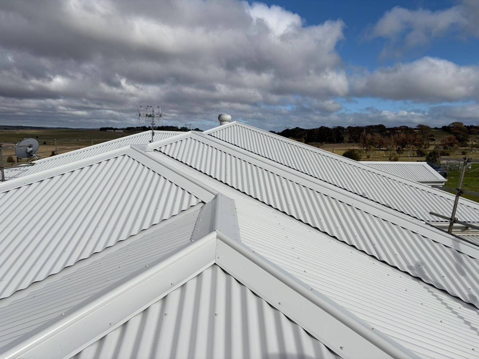 Ridge and hip capping meeting on the same pale corrugated roof, with open farmland behind